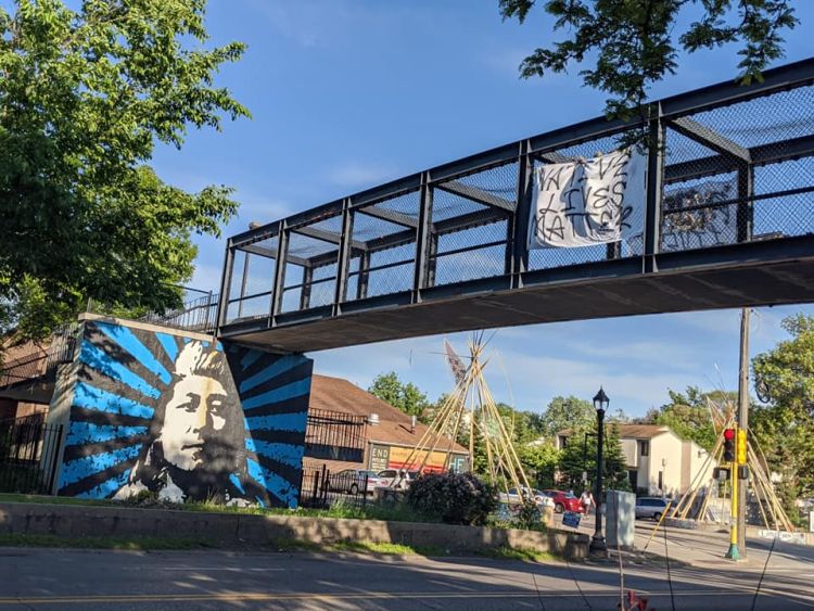 mural of native man with headress appears on concrete wall beneath bridge with protest sign on it, reading "native lives matter"