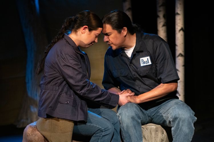 two actors sit together on a stage holding hands and touching foreheads