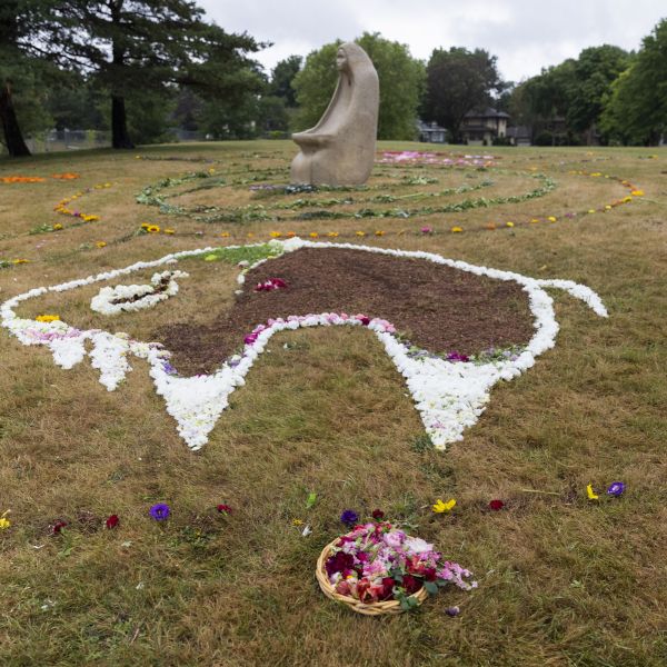 A buffalo drawn in flowers on the ground of a park, with a stone statue of a woman in the background. 