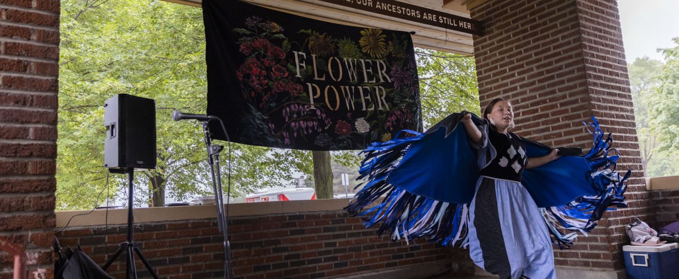 A women in traditional Native American clothing dancing with a sign behind her that says "Flower Power"
