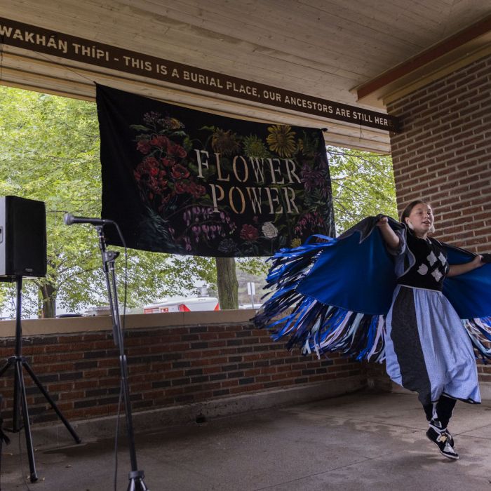A women in traditional Native American clothing dancing with a sign behind her that says "Flower Power"
